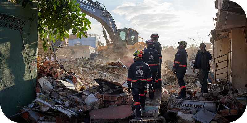 Disaster Assistance Response team members for the Türkiye earthquake crisis response watching an earth moving machine dig through rubble. 